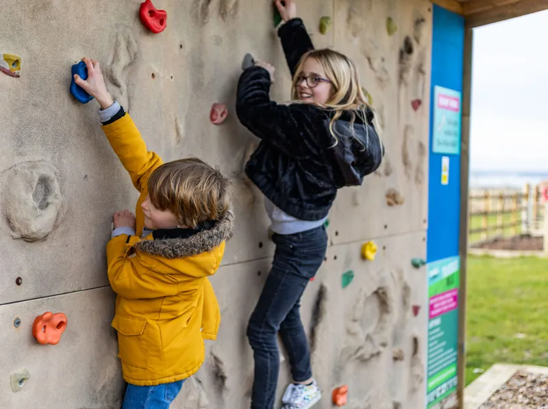 Two children climbing an outdoor rock wall at a farm park in winter.