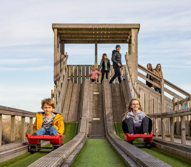 Children riding red sleds down a wooden slide at a farm park playground on a winter day