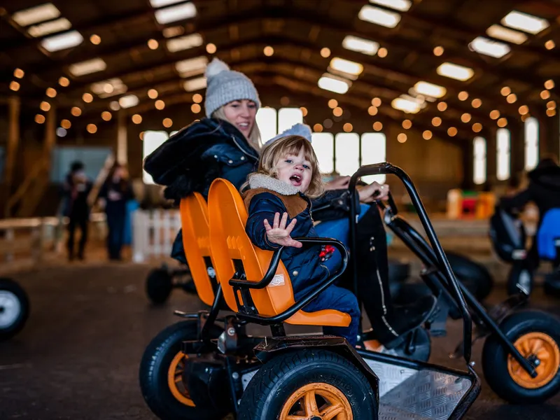 Woman and child riding a go-kart indoors, with the child waving from an orange seat