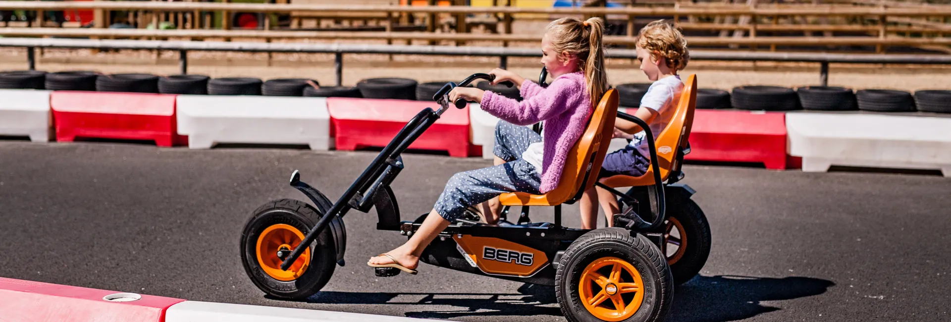 Two children riding a pedal go-kart on an outdoor track with red and white safety barriers