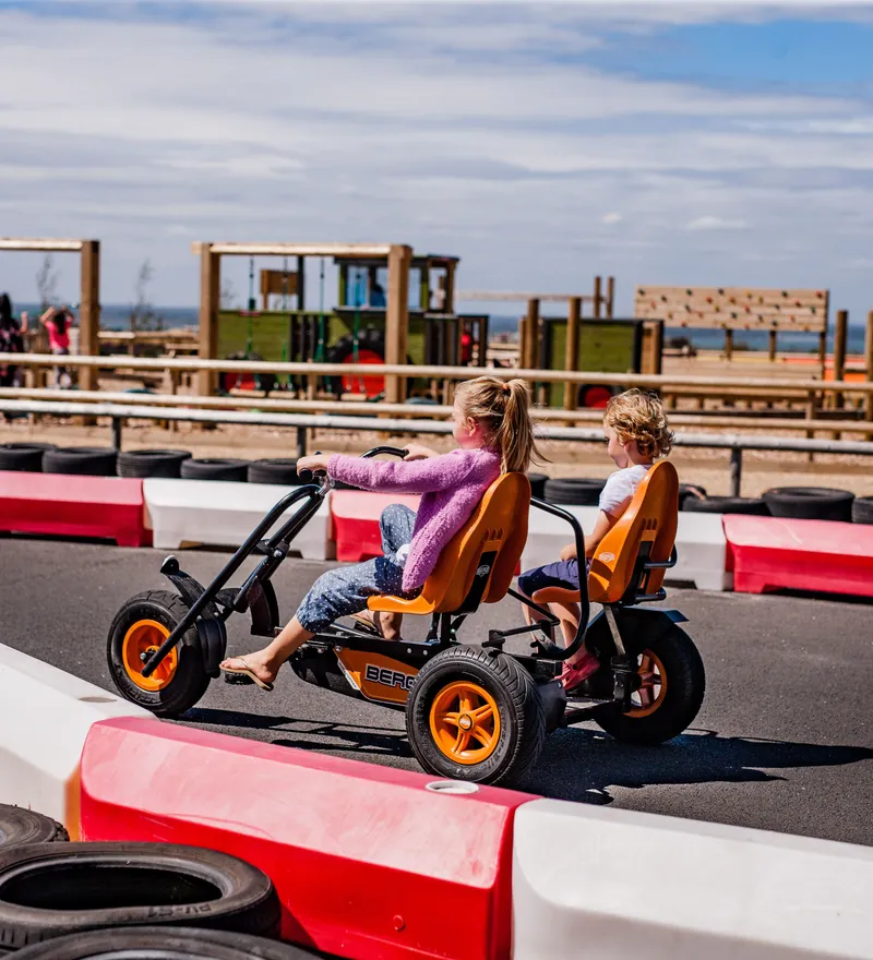 Two children riding a pedal go-kart on an outdoor track with red and white barriers