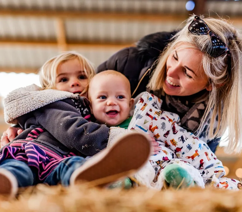 Woman hugging two young children while sitting on hay indoors
