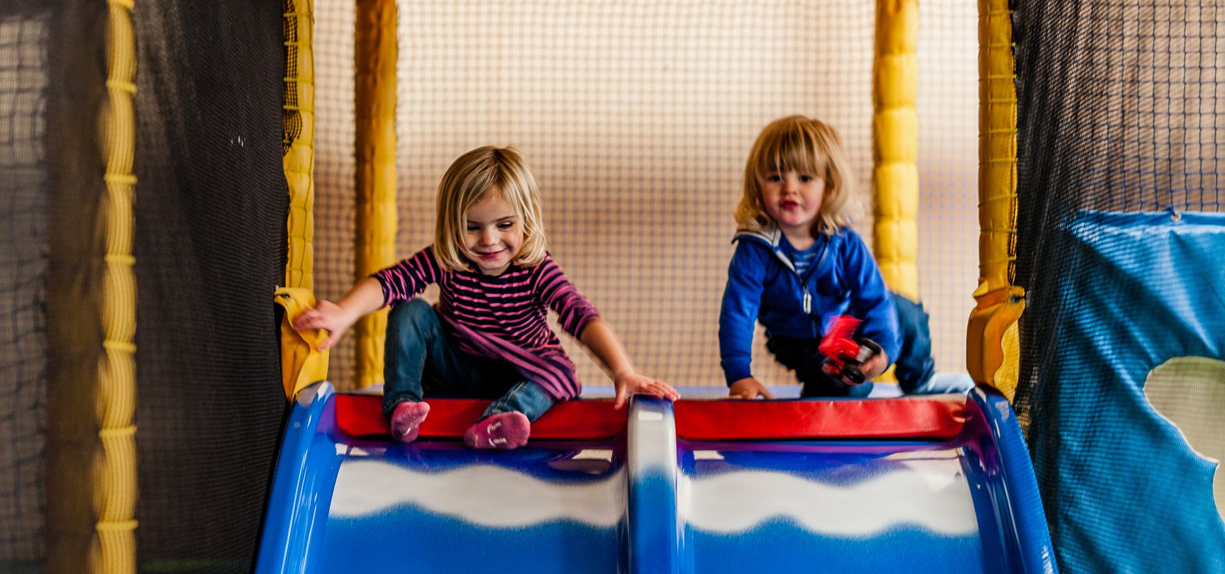 Two young children playing on a double slide in a soft play area