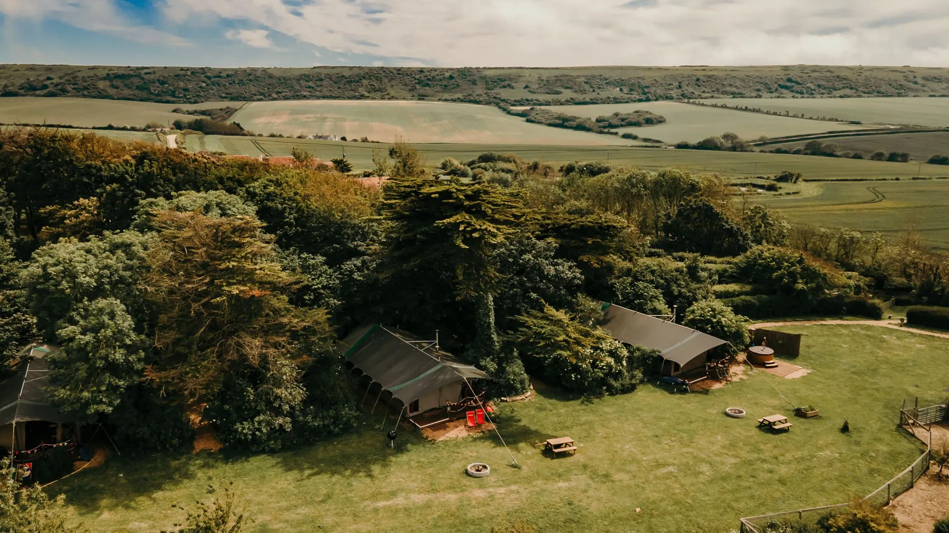 Aerial view of glamping tents beside trees on a grassy field with rolling farmland in the background