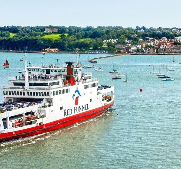 Red Funnel car ferry sailing across a coastal harbour with moored boats and a wooded shoreline in the background