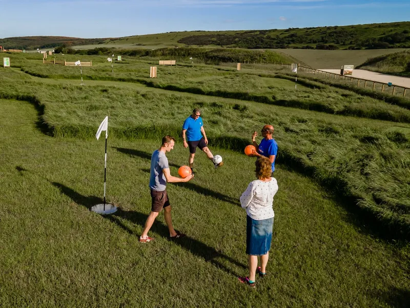 Four adults playing footgolf on a grassy course with a flag, dunes and hills in the background