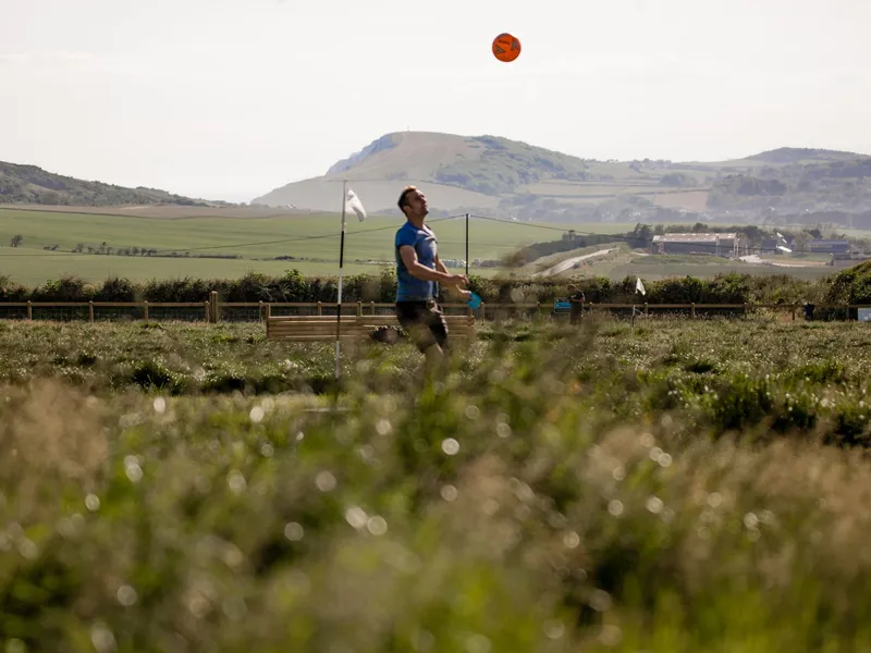 Man kicking an orange football in a grassy field with rolling hills in the background