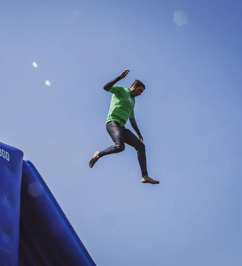 Man in a green top jumps off a large blue inflatable while two people sit on the edge under a clear sky.