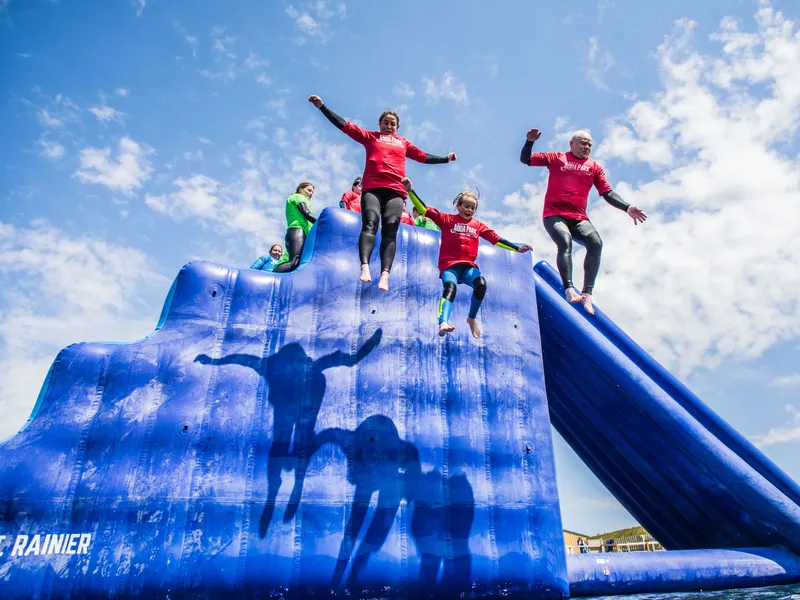 People in red tops jump from a large blue inflatable water slide under a bright, partly cloudy sky