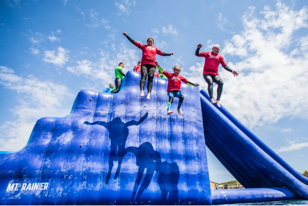 People in red tops jump from a large blue inflatable water slide under a bright, partly cloudy sky