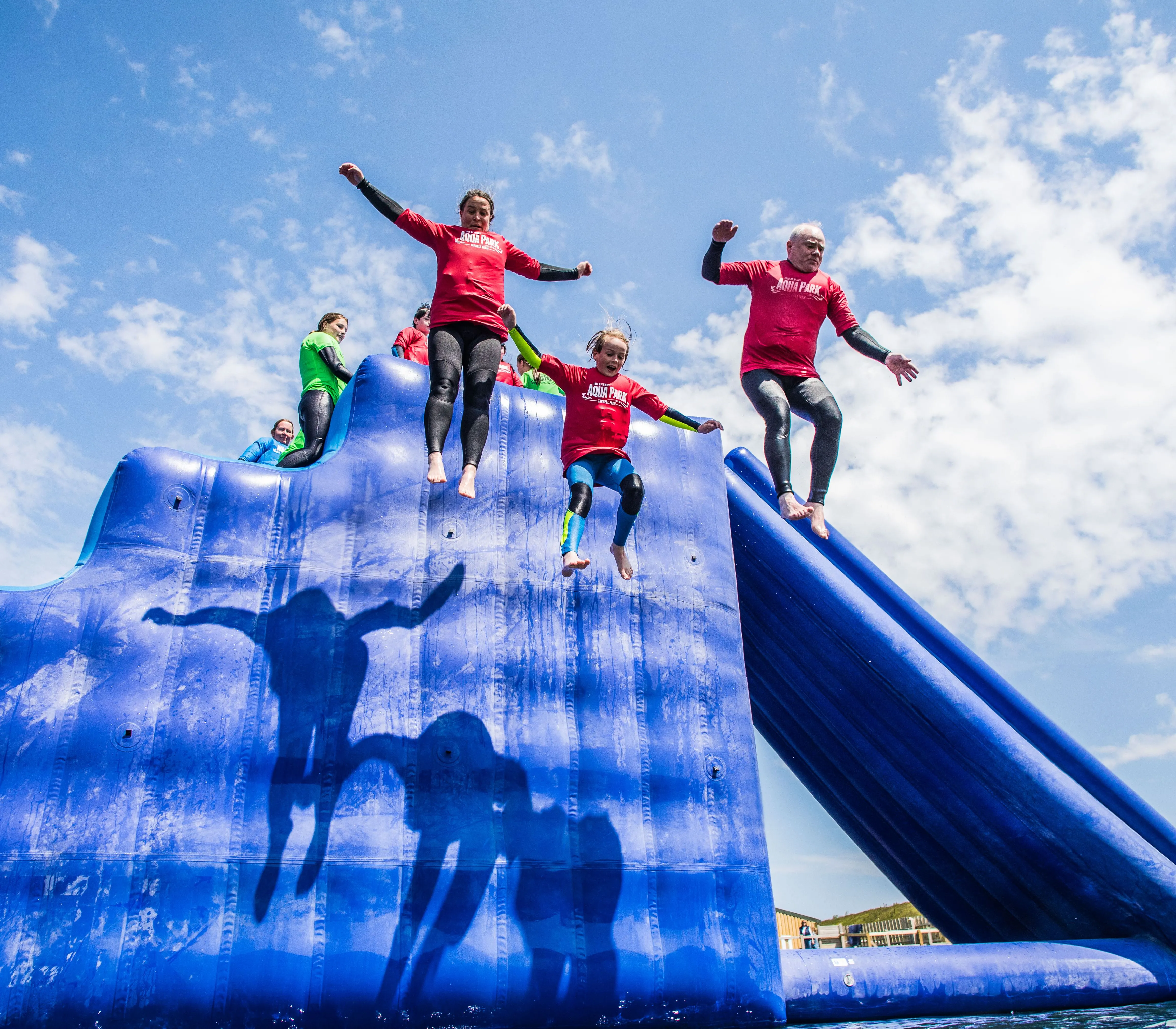 People in red tops jump from a large blue inflatable water slide under a bright, partly cloudy sky