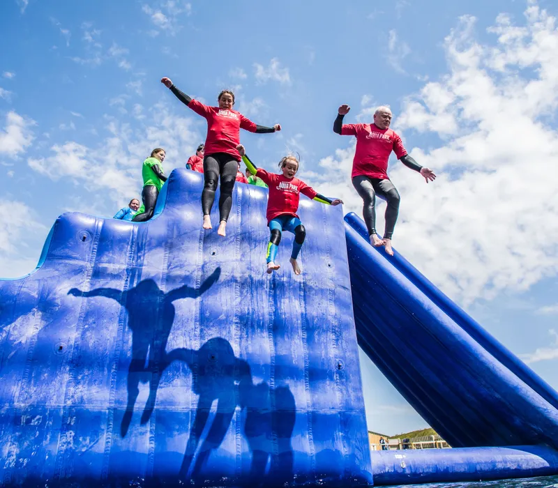 People in red tops jump from a large blue inflatable water slide under a bright, partly cloudy sky