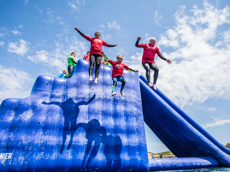 People in red tops jump from a large blue inflatable water slide under a bright, partly cloudy sky