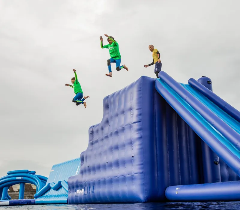 People jumping off a large blue inflatable water slide into the water