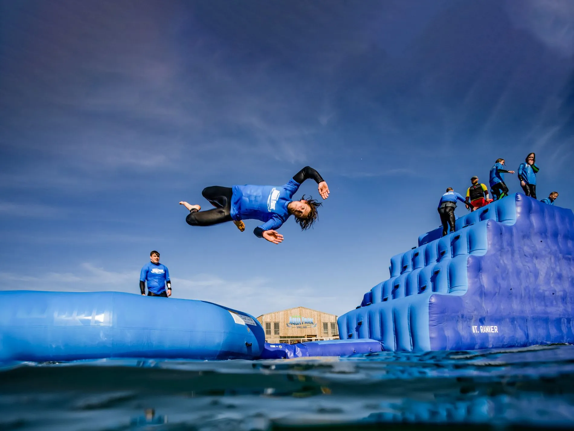 Person in a blue wetsuit doing a mid-air flip above an inflatable water park obstacle