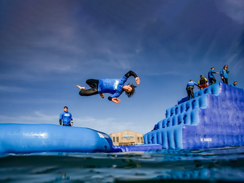 Person in a blue wetsuit doing a mid-air flip above an inflatable water park obstacle