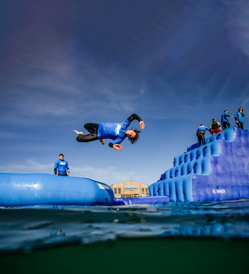 Person in a blue wetsuit doing a mid-air flip above an inflatable water park obstacle