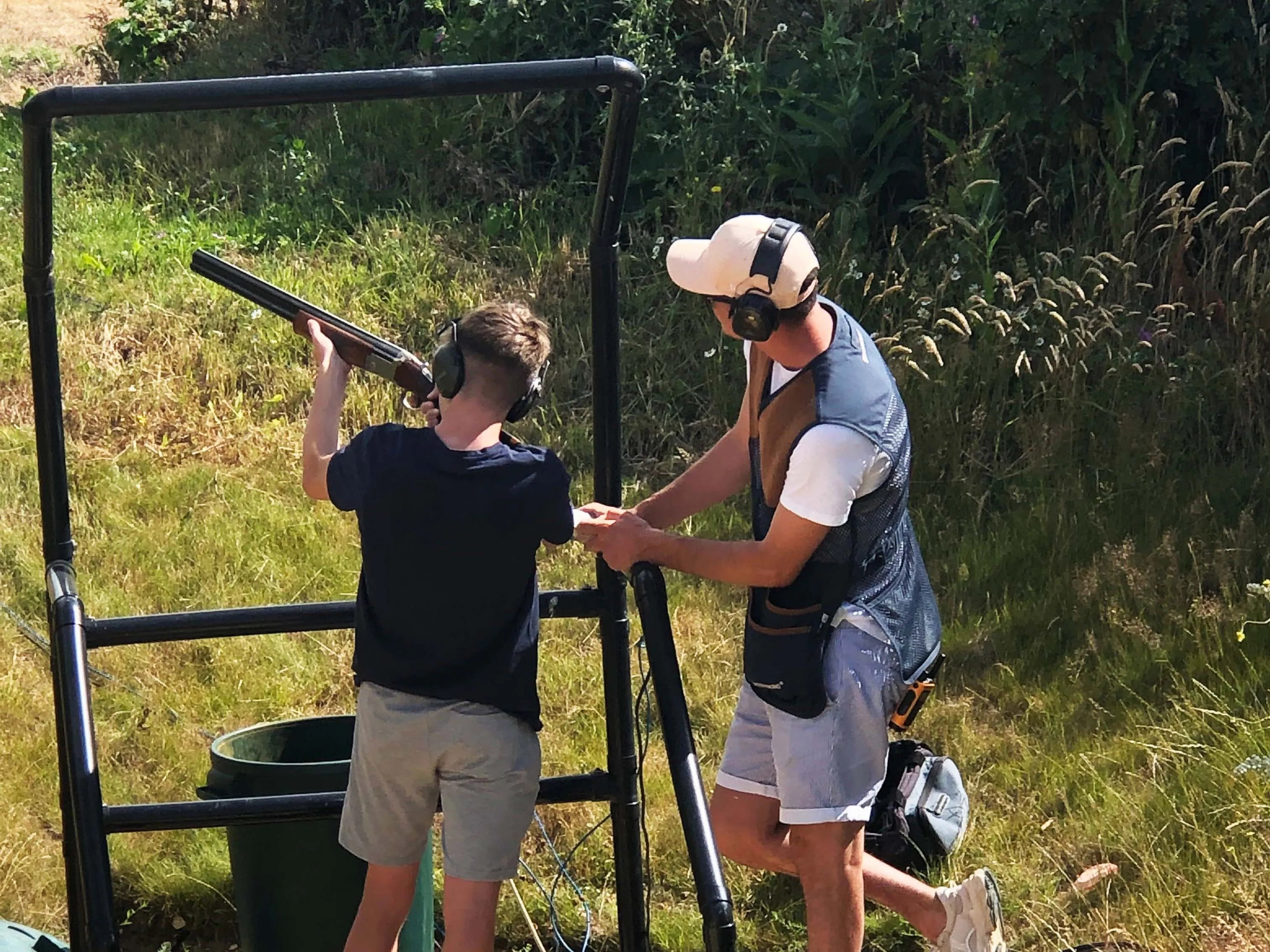 Young shooter aiming a shotgun at a clay pigeon shooting stand while an instructor assists, both wearing ear defenders outdoors.