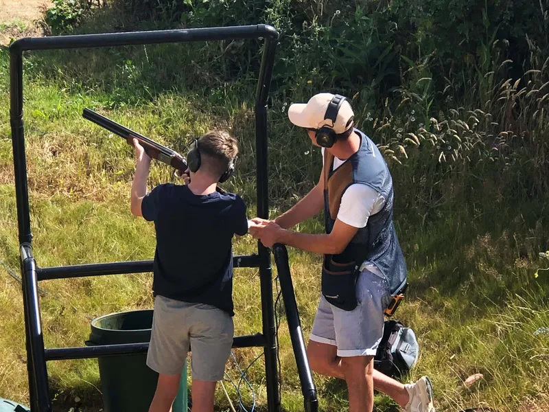 Young shooter aiming a shotgun at a clay pigeon shooting stand while an instructor assists, both wearing ear defenders outdoors.