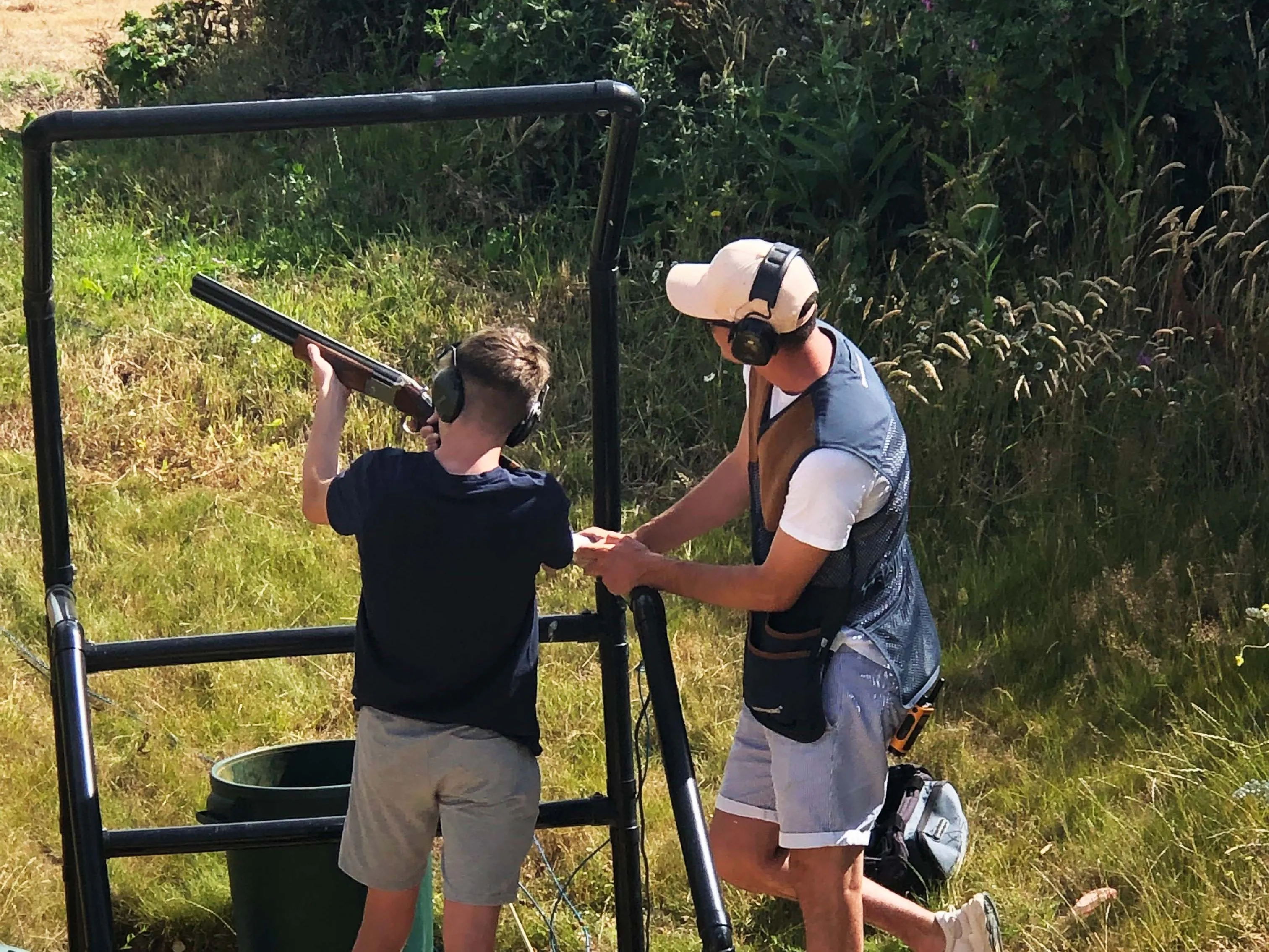 Young shooter aiming a shotgun at a clay pigeon shooting stand while an instructor assists, both wearing ear defenders outdoors.