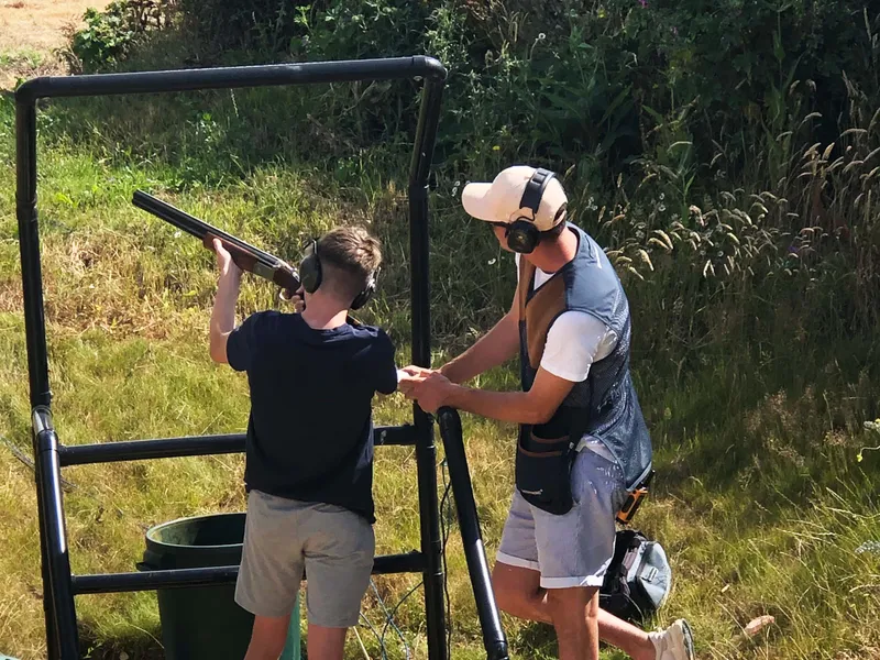 Young shooter aiming a shotgun at a clay pigeon shooting stand while an instructor assists, both wearing ear defenders outdoors.