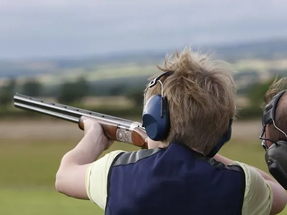Person wearing ear defenders aiming a shotgun outdoors at a shooting range
