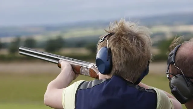 Person wearing ear defenders aiming a shotgun outdoors at a shooting range