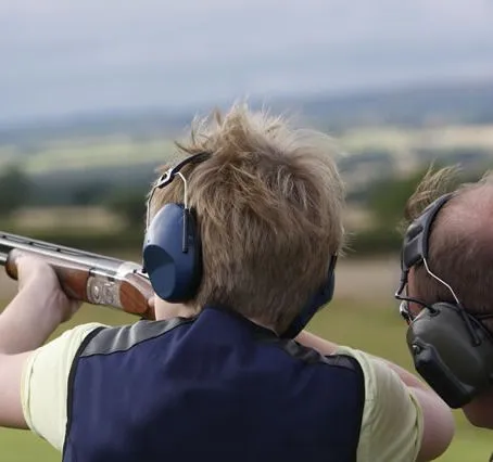 Person wearing ear defenders aiming a shotgun outdoors at a shooting range