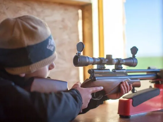 Child aiming a scoped rifle from a shooting bench in a wooden hide