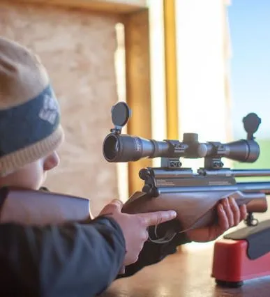 Child aiming a scoped rifle from a shooting bench in a wooden hide