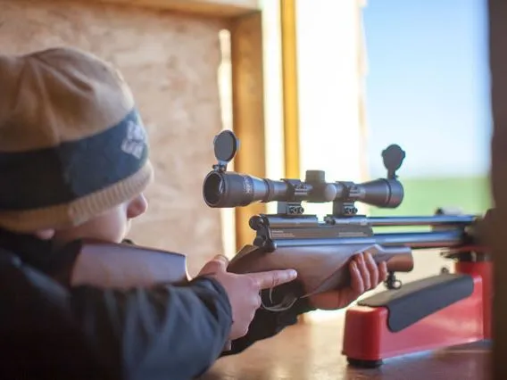 Child aiming a scoped rifle from a shooting bench in a wooden hide