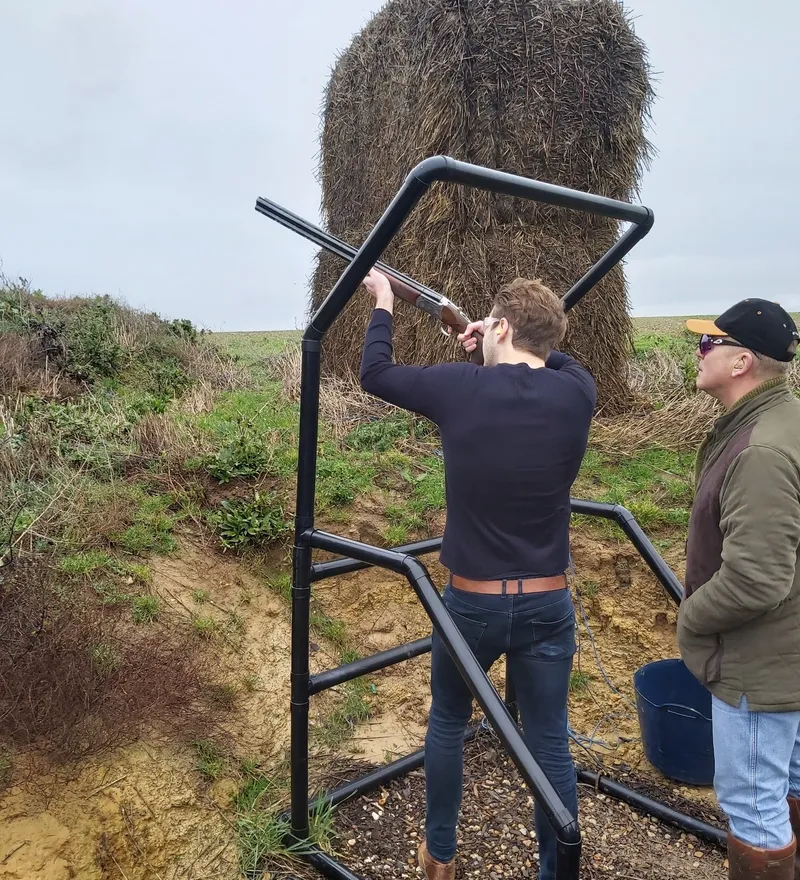 Man aiming a shotgun at an outdoor clay shooting stand with a large hay bale behind, while another man watches