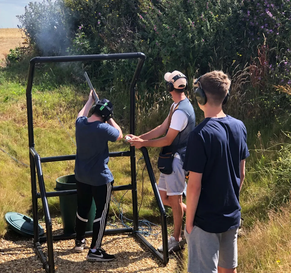 Three people wearing ear defenders at an outdoor clay pigeon shooting stand, with one aiming a shotgun