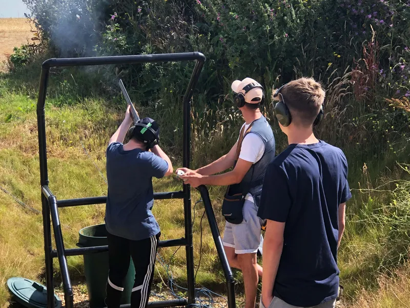 Three people wearing ear defenders at an outdoor clay pigeon shooting stand, with one aiming a shotgun