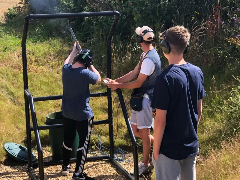 Three people wearing ear defenders at an outdoor clay pigeon shooting stand, with one aiming a shotgun