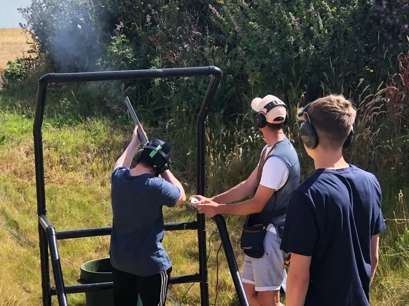 Three people wearing ear defenders at an outdoor clay pigeon shooting stand, with one aiming a shotgun