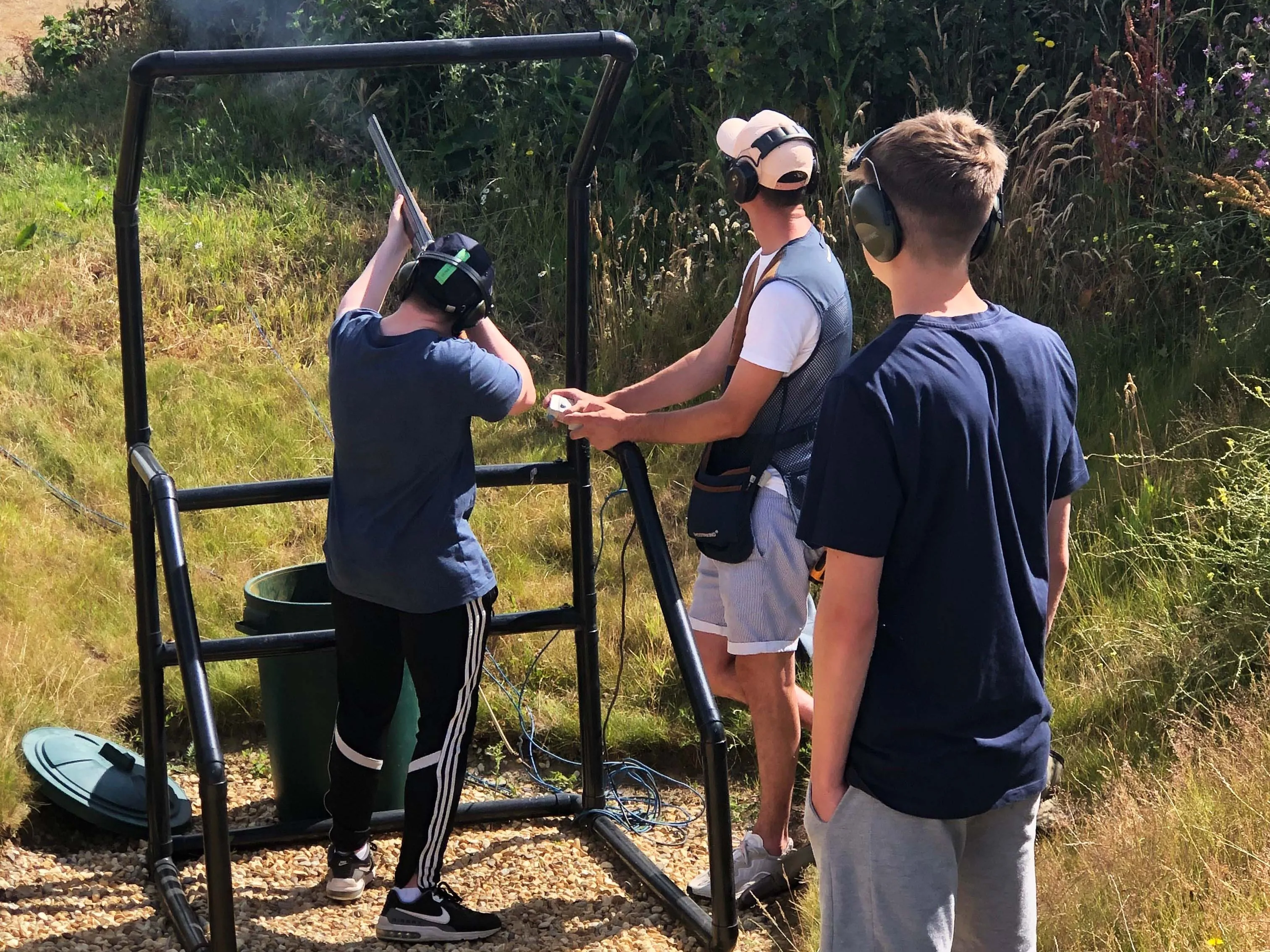 Three people wearing ear defenders at an outdoor clay pigeon shooting stand, with one aiming a shotgun