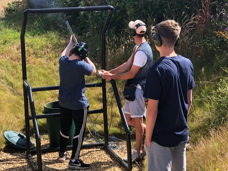 Three people wearing ear defenders at an outdoor clay pigeon shooting stand, with one aiming a shotgun
