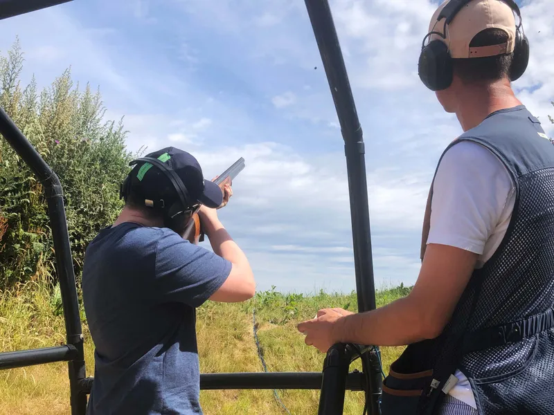 Person aiming a shotgun at a clay pigeon shooting range while an instructor watches