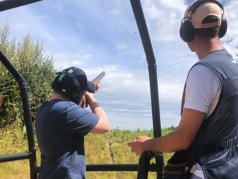 Person aiming a shotgun at a clay pigeon shooting range while an instructor watches