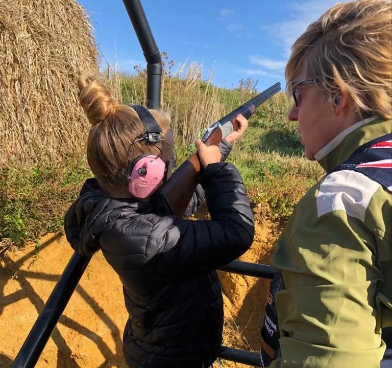 Person wearing ear defenders aiming a shotgun at an outdoor clay shooting range with an instructor beside them