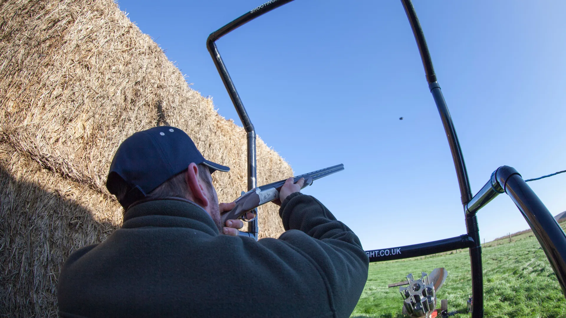 Man aiming a shotgun at a clay pigeon on an outdoor shooting range beside hay bales