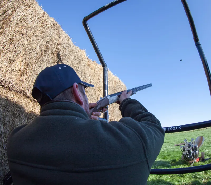 Man aiming a shotgun at a clay pigeon on an outdoor shooting range beside hay bales