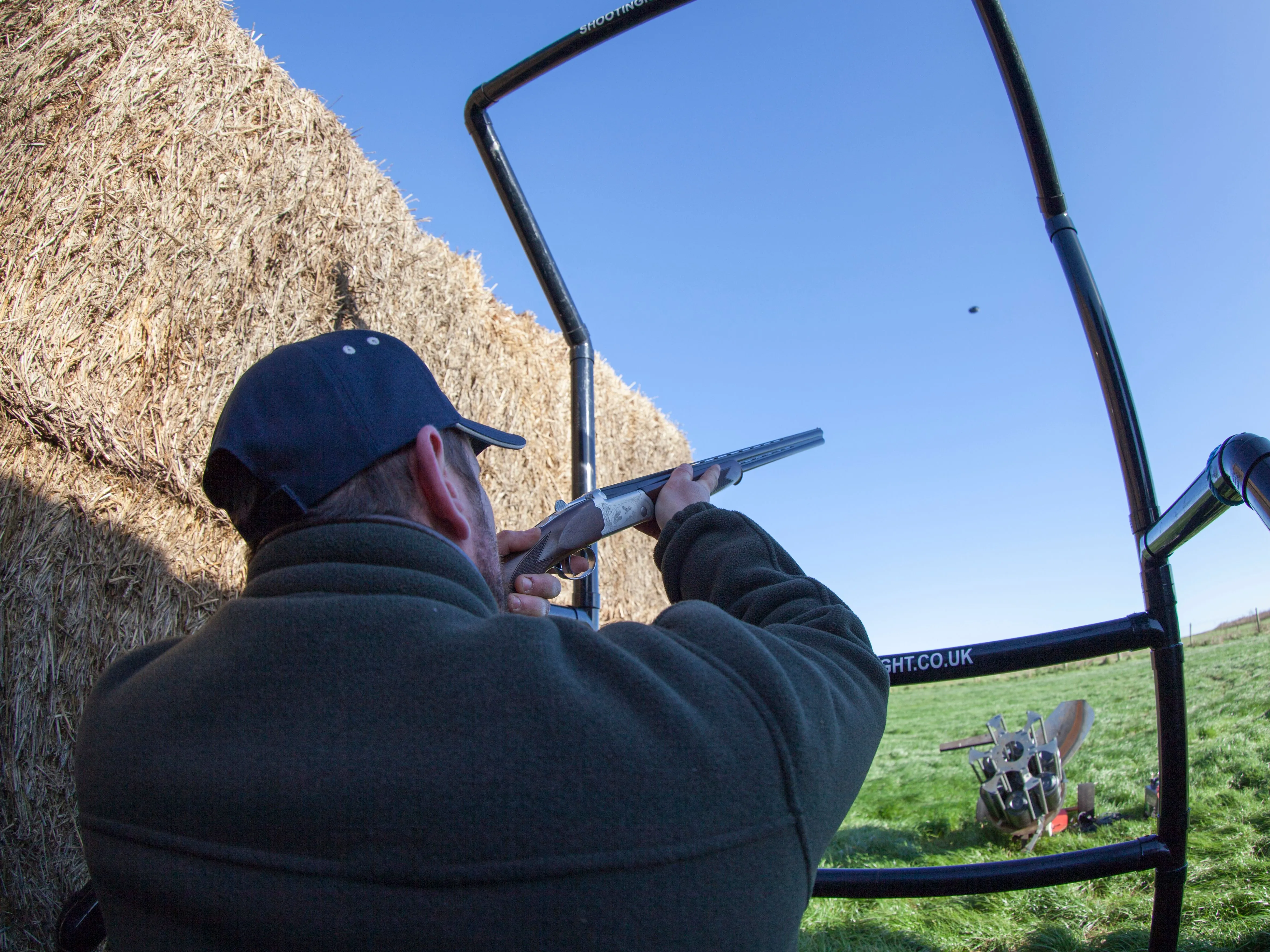 Man aiming a shotgun at a clay pigeon on an outdoor shooting range beside hay bales