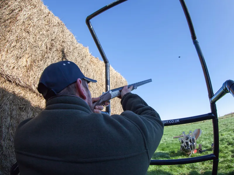 Man aiming a shotgun at a clay pigeon on an outdoor shooting range beside hay bales