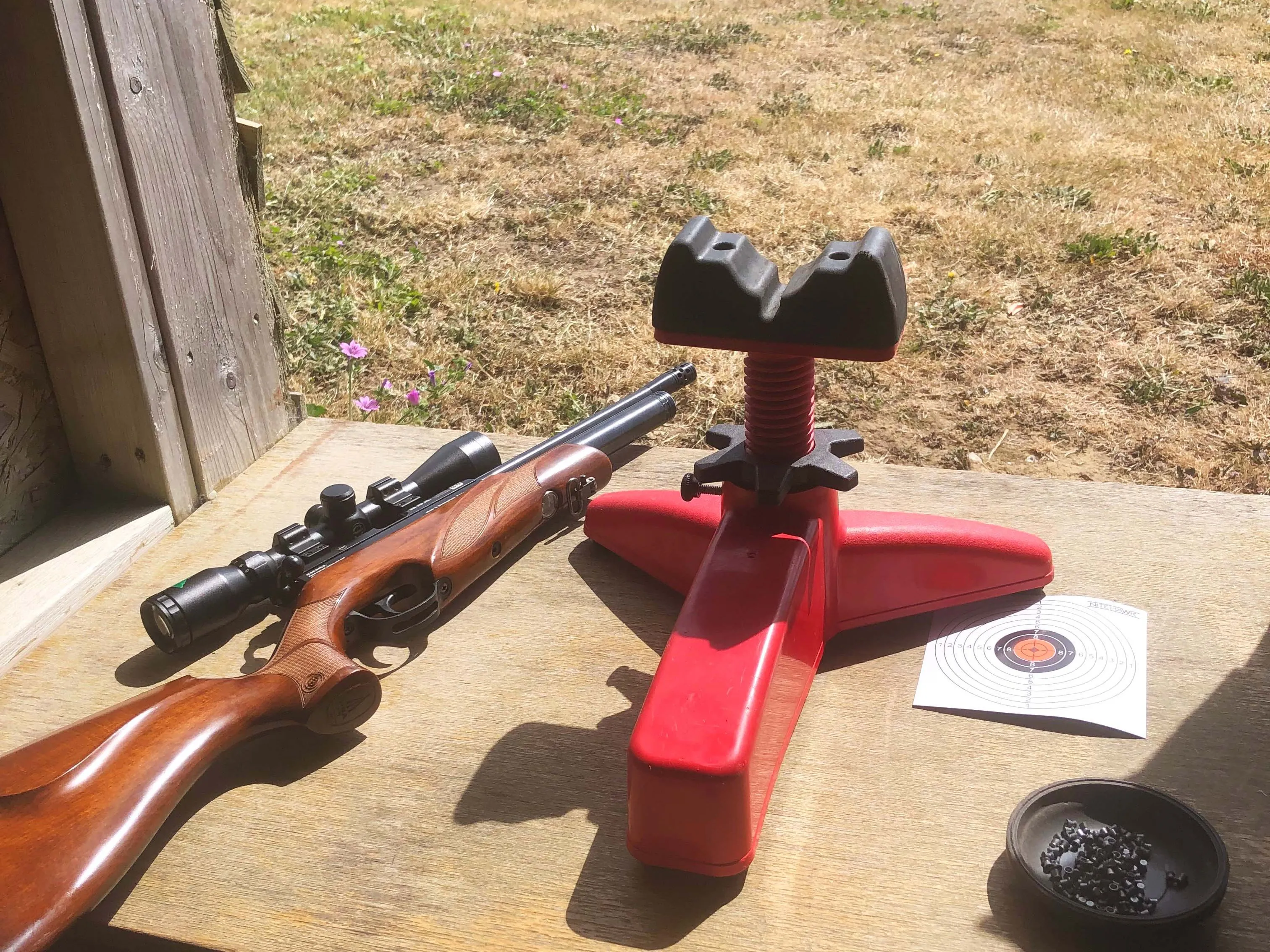 Rifle with scope beside a red shooting rest on a wooden bench at an outdoor range