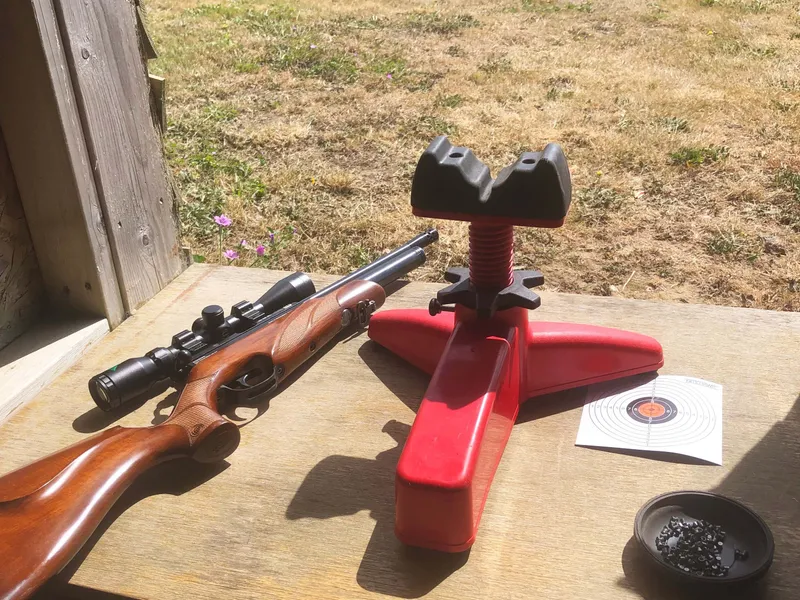Rifle with scope beside a red shooting rest on a wooden bench at an outdoor range