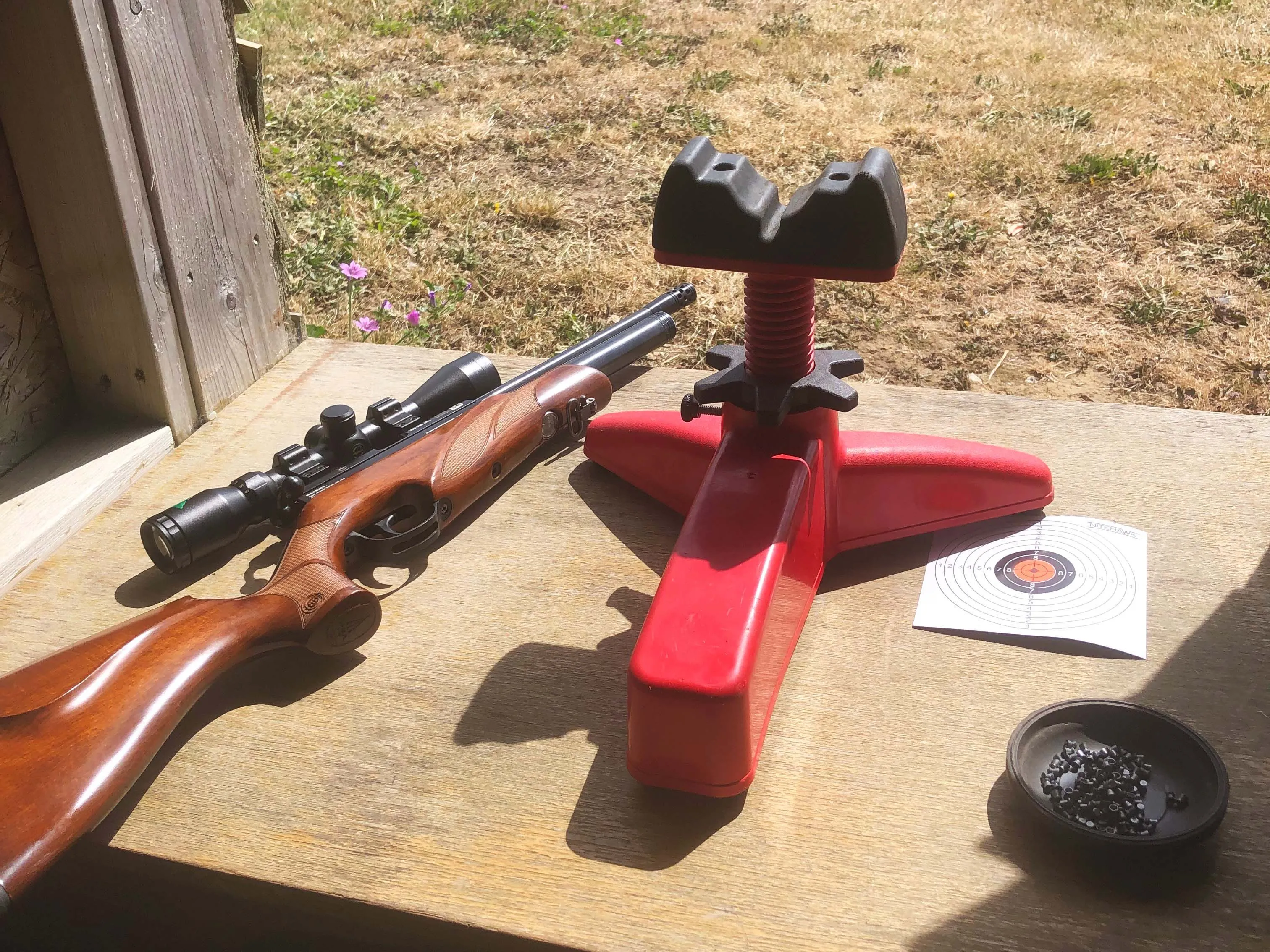 Rifle with scope beside a red shooting rest on a wooden bench at an outdoor range