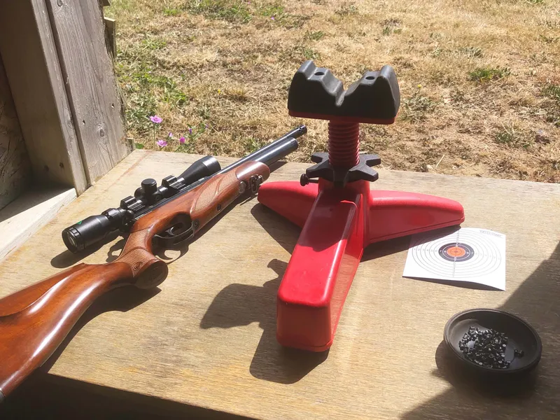 Rifle with scope beside a red shooting rest on a wooden bench at an outdoor range