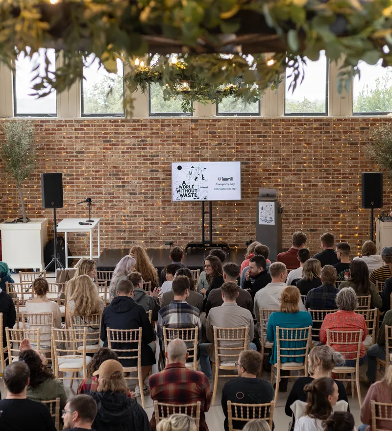 Audience seated in a bright event hall facing a small stage with a presentation screen and speakers